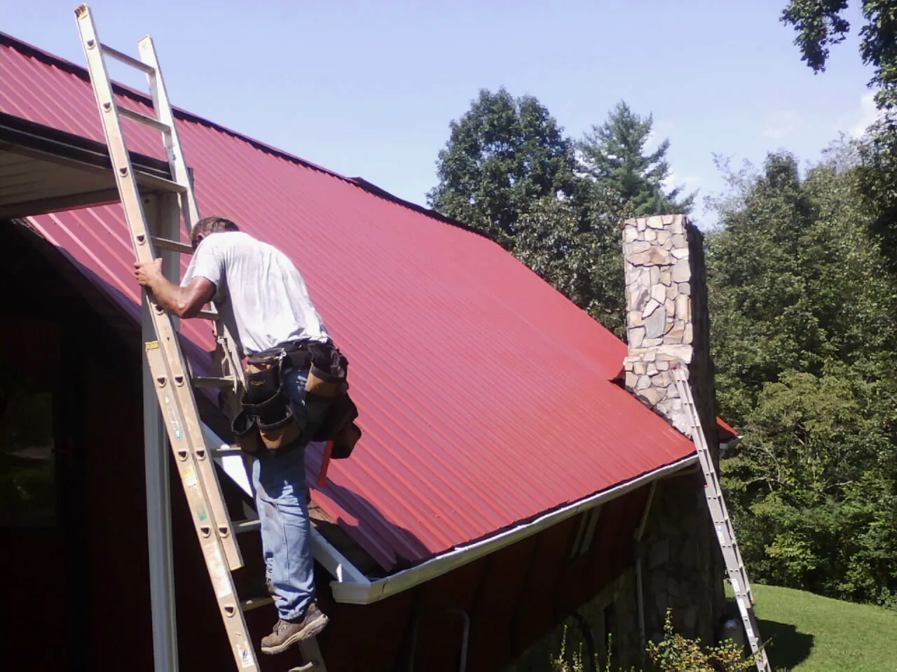 Roofer on a ladder beside a red metal roof and stone chimney — Soffit & Fascia Repair by Roofing Brothers in Sewickley
